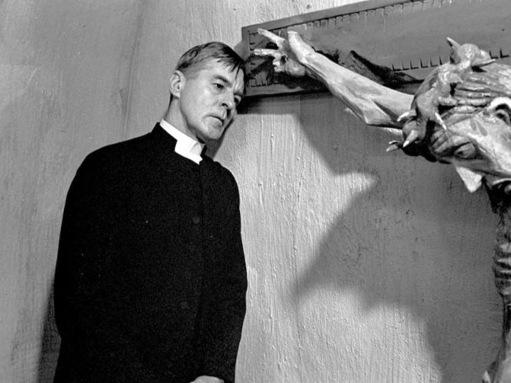 Black and white still from the film "Winter Light". It features a priest staring forlornly next to a crucifixion of Christ.