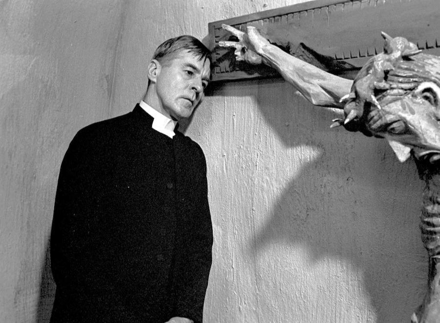 Black and white still from the film "Winter Light". It features a priest staring forlornly next to a crucifixion of Christ.