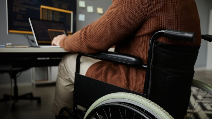 Image of a person in a wheelchair in front of a laptop. The person's head is not shown, they are wearing a brown sweater with khaki pants. The wheelchair is black.