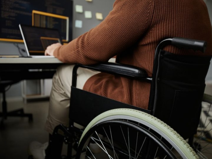 Image of a person in a wheelchair in front of a laptop. The person's head is not shown, they are wearing a brown sweater with khaki pants. The wheelchair is black.