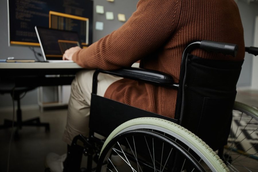 Image of a person in a wheelchair in front of a laptop. The person's head is not shown, they are wearing a brown sweater with khaki pants. The wheelchair is black.