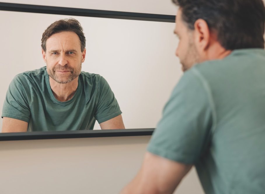 A man in a teal T-shirt, Tarshis, studies himself in the mirror