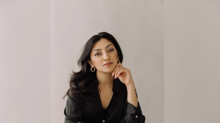 A woman in a blazer sits at a table, looking into the camera, against a neutral-coloured background.