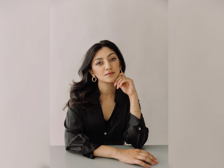 A woman in a blazer sits at a table, looking into the camera, against a neutral-coloured background.