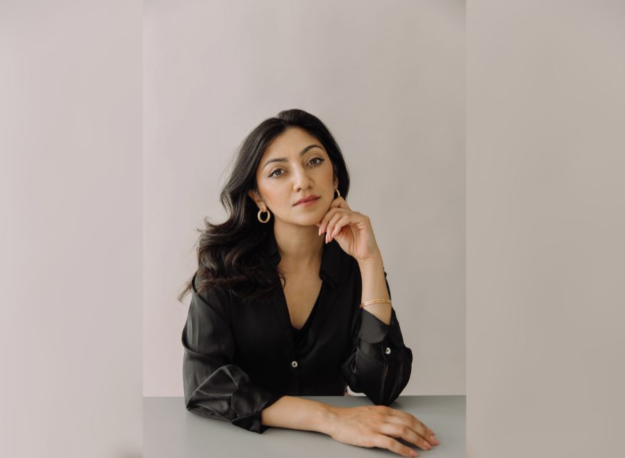 A woman in a blazer sits at a table, looking into the camera, against a neutral-coloured background.