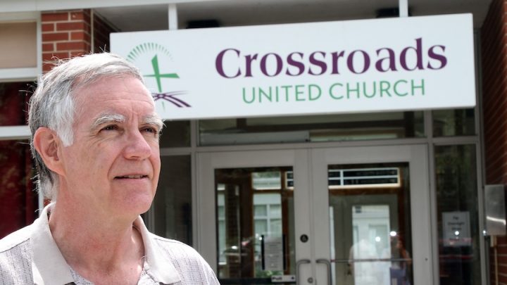 An older white man with white hair and in a brown button-up shirt stands in front of a building with double doors. There is a sign that says, "Crossroads United Church."