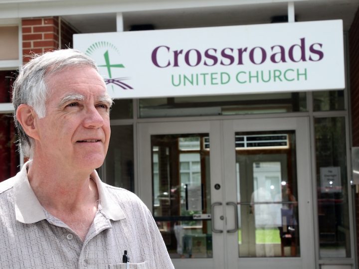 An older white man with white hair and in a brown button-up shirt stands in front of a building with double doors. There is a sign that says, "Crossroads United Church."