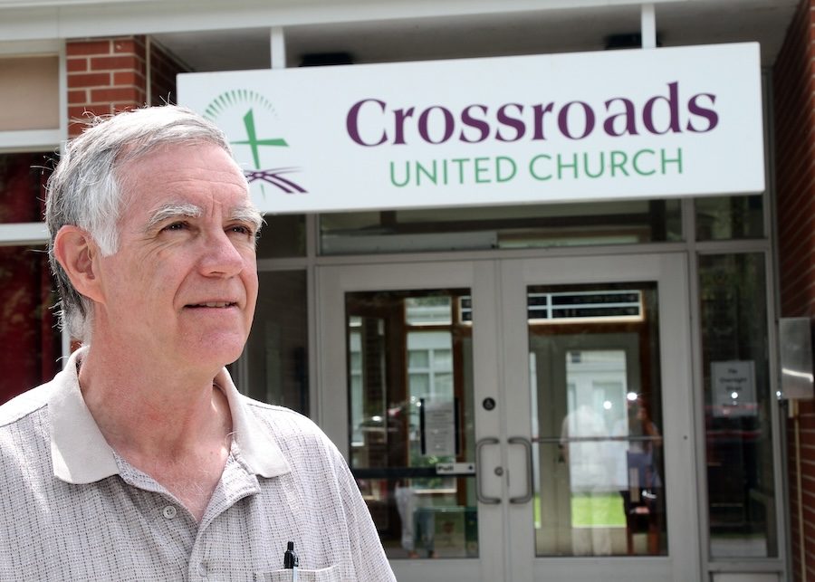 An older white man with white hair and in a brown button-up shirt stands in front of a building with double doors. There is a sign that says, "Crossroads United Church."