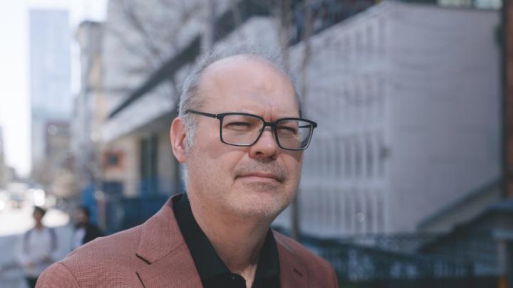 A middle-aged white man who is balding and wearing black-framed glasses, a black button-up shirt and a brown blazer poses in front of a building.