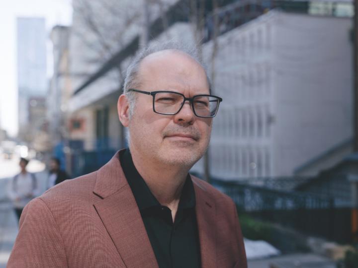 A middle-aged white man who is balding and wearing black-framed glasses, a black button-up shirt and a brown blazer poses in front of a building.