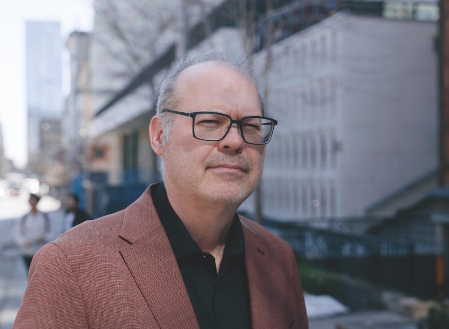 A middle-aged white man who is balding and wearing black-framed glasses, a black button-up shirt and a brown blazer poses in front of a building.