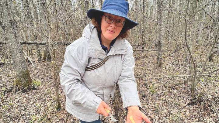 A white woman with curly brown hair stands in a forest with her hand outstretched. In her left hand is a brown mushroom. In her right hand is a small knife. She is wearing a navy bucket hat, light grey jacket, blue jeans and black boots. The background is a forest with trees and the ground is full of dead leaves and logs.