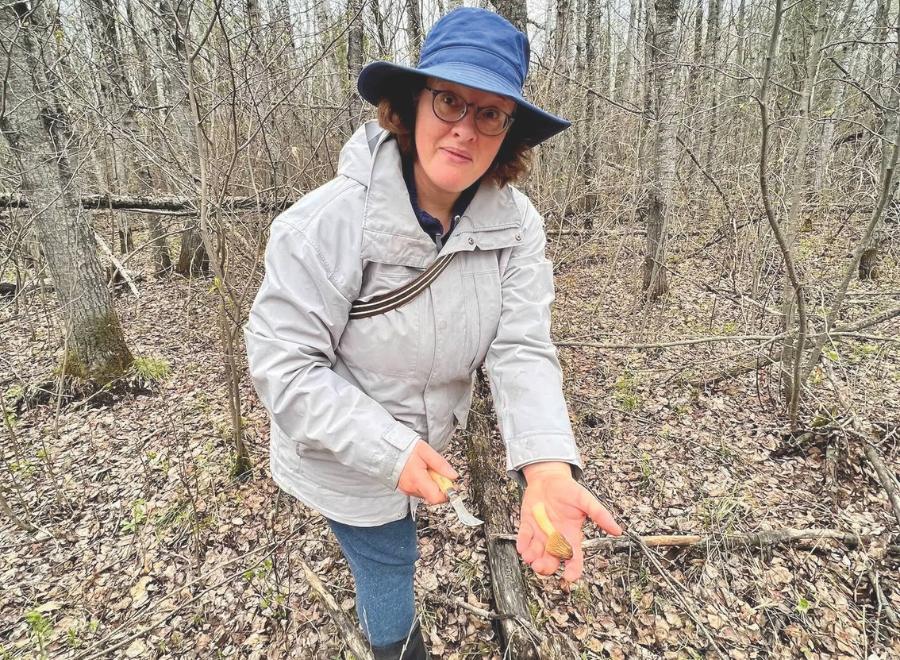 A white woman with curly brown hair stands in a forest with her hand outstretched. In her left hand is a brown mushroom. In her right hand is a small knife. She is wearing a navy bucket hat, light grey jacket, blue jeans and black boots. The background is a forest with trees and the ground is full of dead leaves and logs.