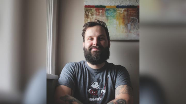 A white male with brown hair and a brown beard sits in front of a painting. He is smiling and wearing a short sleeve t-shirt with Mickey Mouse on it.