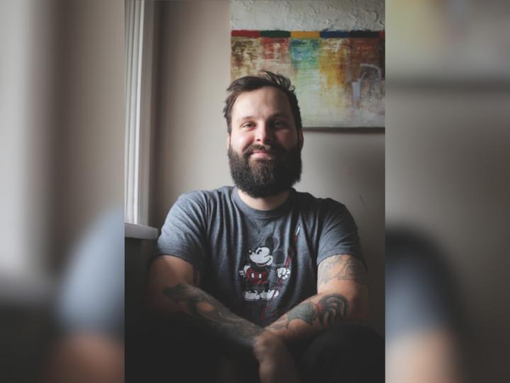 A white male with brown hair and a brown beard sits in front of a painting. He is smiling and wearing a short sleeve t-shirt with Mickey Mouse on it.