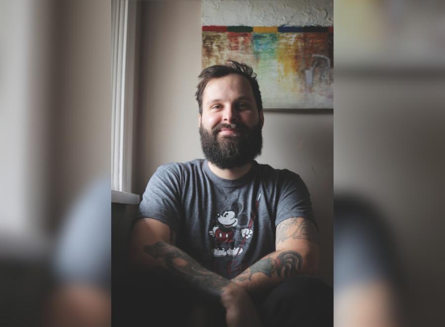 A white male with brown hair and a brown beard sits in front of a painting. He is smiling and wearing a short sleeve t-shirt with Mickey Mouse on it.