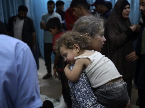 A child covered in dust is carrying another smaller child covered in dust. The younger child is wearing a dirty white tank top and black shorts. They both have curly brown hair that is covered in dust. In the background are adults including one woman in a black hijab.