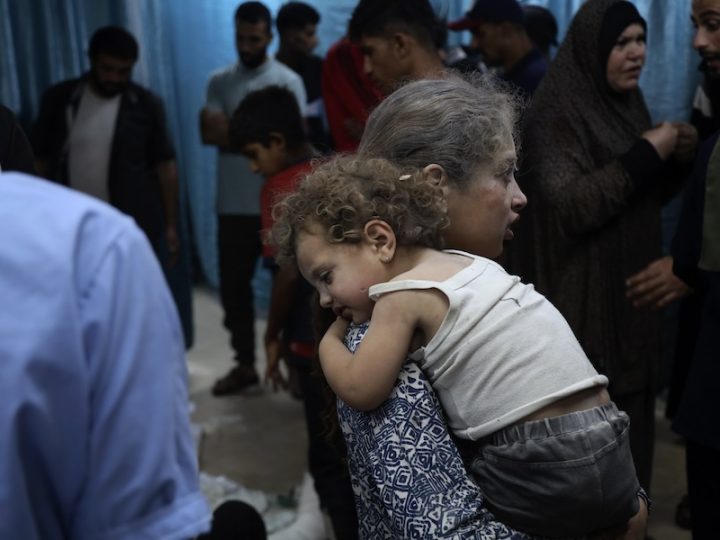 A child covered in dust is carrying another smaller child covered in dust. The younger child is wearing a dirty white tank top and black shorts. They both have curly brown hair that is covered in dust. In the background are adults including one woman in a black hijab.