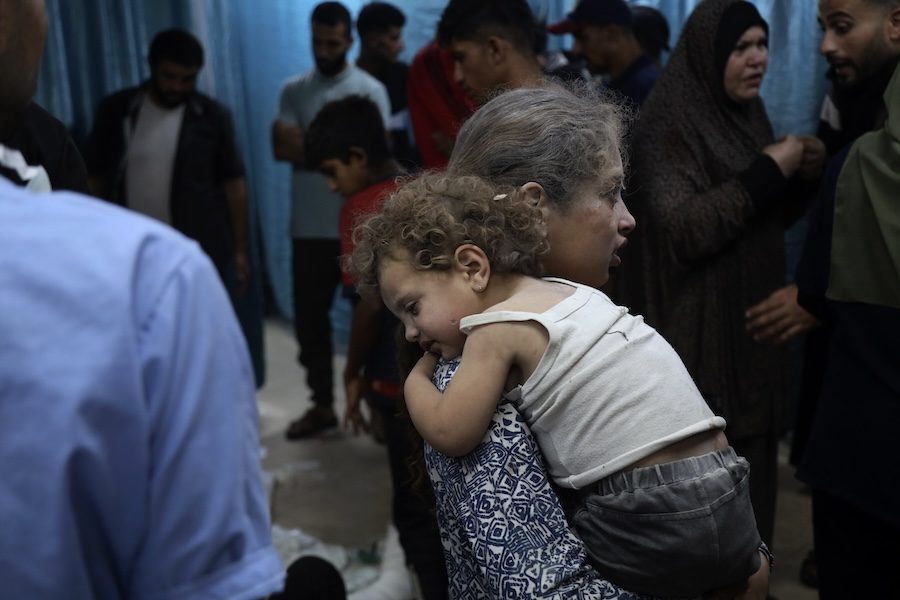 A child covered in dust is carrying another smaller child covered in dust. The younger child is wearing a dirty white tank top and black shorts. They both have curly brown hair that is covered in dust. In the background are adults including one woman in a black hijab.