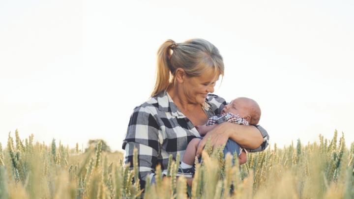 An older white woman with grey hair in a ponytail holds a baby in her arms. She is wearing a black and white flannel shirt and is standing in a field of corn.