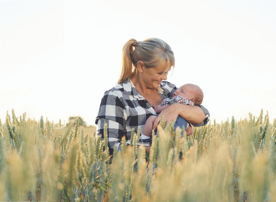 An older white woman with grey hair in a ponytail holds a baby in her arms. She is wearing a black and white flannel shirt and is standing in a field of corn.