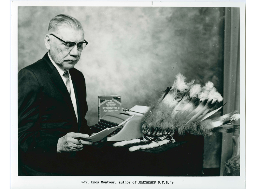 Black and white image of a middle-aged man in glasses and a dark suit and tie. He is holding paper in his head. Next to him is a table with a headdress, typewriter and a book.