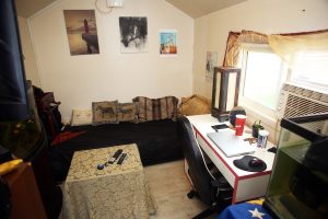 Interior of a tiny cabin featuring a sleeping area with cushions, photos on the wall, a small coffee table with keys, two black remote controls. There is a white desk on the side with a silver laptop on it, a black desk chair tucked in, a black mouse and mouse pad, a lantern, a red Tim Horton's cup, a plant, a mirror and brown beads on the desk. It is next to a window that has an a/c unit and the sun is shining in through the window lighting up the small room. 