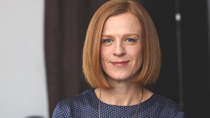 Headshot of a white woman with short-red hair that is parted on the side. She is smiling and wearing a purple shirt and bronze necklace