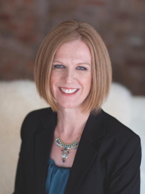 Headshot of a white woman with a short red bob. She is smiling and wearing a blue shirt, blue necklace and black blazer.