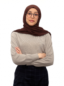 Headshot of a brown-skinned woman in a maroon hijab. She is wearing black-framed glasses, a beige long-sleeve shirt and black pants. Her arms are crossed in front of her chest. 