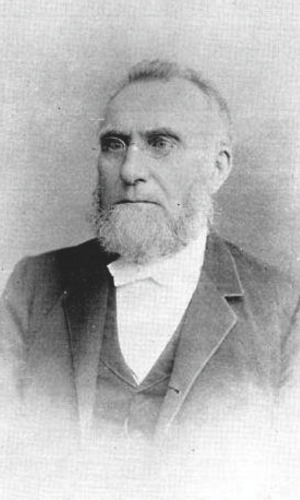 Black and white photo of a white man with small glasses, beard and he is wearing a suit jacket and white shirt with a bow. 