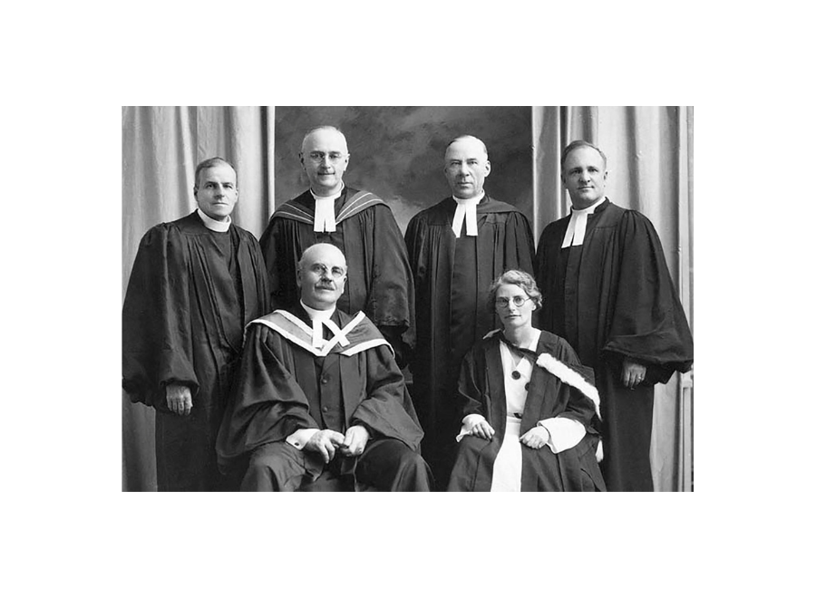 Black and white photo of six white people in reverend clothing. The front row has three people seated. They are all wearing black robes and white collars. On the front row, right, is a woman wearing glasses. 