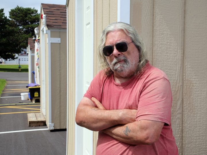 A white middle-aged man stands in front of a tiny cabin. He has long white hair, a white beard, black sunglasses on and is wearing a pink shirt. His hands are crossed in front of his chest.