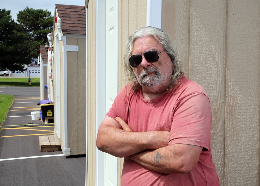 A white middle-aged man stands in front of a tiny cabin. He has long white hair, a white beard, black sunglasses on and is wearing a pink shirt. His hands are crossed in front of his chest.