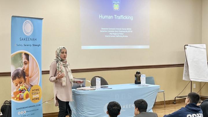 A brown-skinned woman in a hijab is doing a presentation on human trafficking. There is a screen that has a police logo on it and says, "Human trafficking" and she is standing next to it by a table with chairs and a sign that says Sakeenah. There are students sitting on the floor in front of her. You can see the students' backs and they are in blue shirts and have brown hair.