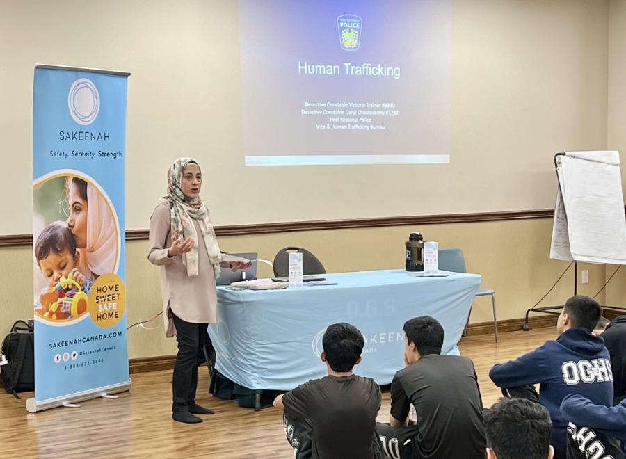 A brown-skinned woman in a hijab is doing a presentation on human trafficking. There is a screen that has a police logo on it and says, "Human trafficking" and she is standing next to it by a table with chairs and a sign that says Sakeenah. There are students sitting on the floor in front of her. You can see the students' backs and they are in blue shirts and have brown hair.