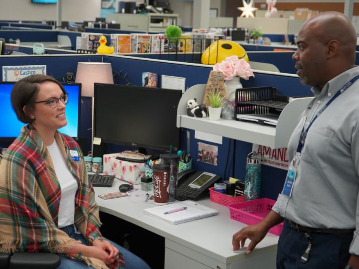 A man stands beside a desk at an office where he speaks with a woman sitting in a desk chair.