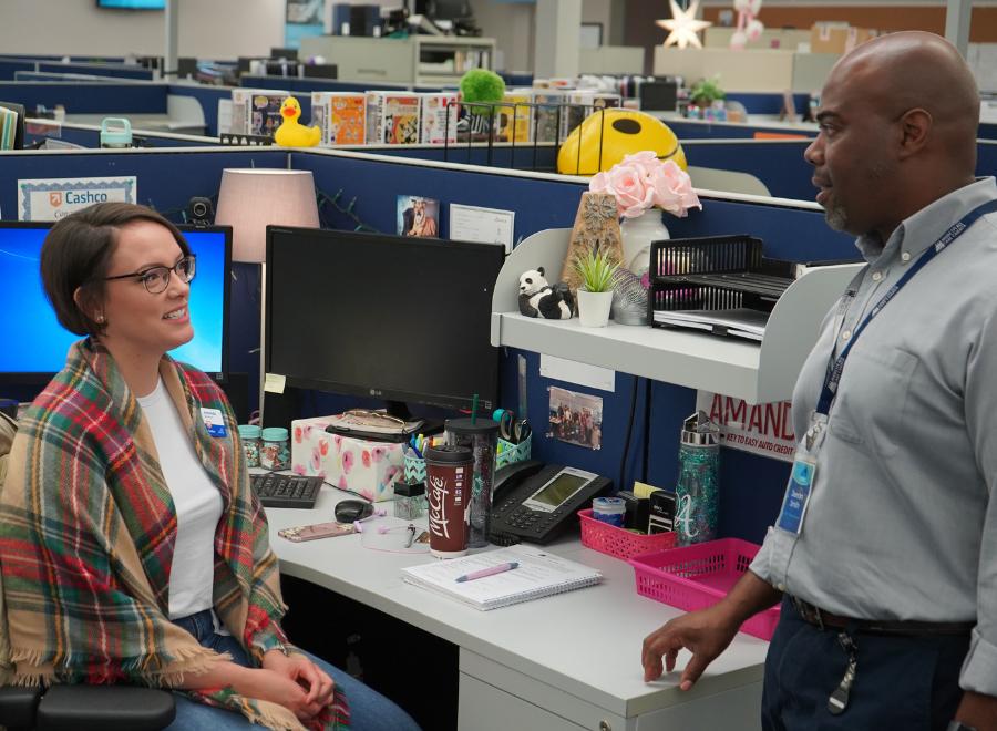 A man stands beside a desk at an office where he speaks with a woman sitting in a desk chair.