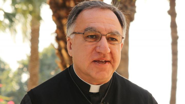 A man dressed in a black shirt with a priest's collar. He wears glasses.