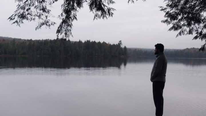 Black and white photo of a man standing by a lake. It is a side profile of the man.