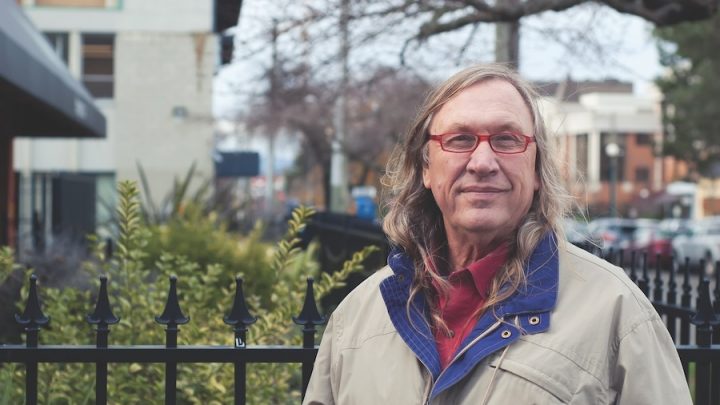 A white middle-aged man stands in front of a black fence and yard. He has long brown-grey hair, wears red-framed glasses, a red shirt and a brown coat with a blue collar.