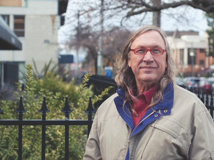 A white middle-aged man stands in front of a black fence and yard. He has long brown-grey hair, wears red-framed glasses, a red shirt and a brown coat with a blue collar.