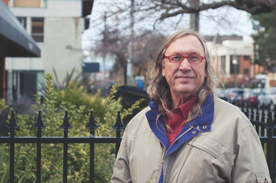 A white middle-aged man stands in front of a black fence and yard. He has long brown-grey hair, wears red-framed glasses, a red shirt and a brown coat with a blue collar.