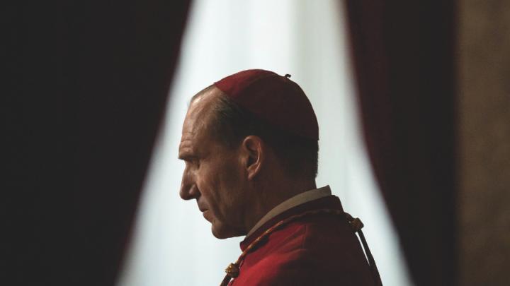 A white man in a red cap and red robe stands in front of a window. The perspective is from his side profile.