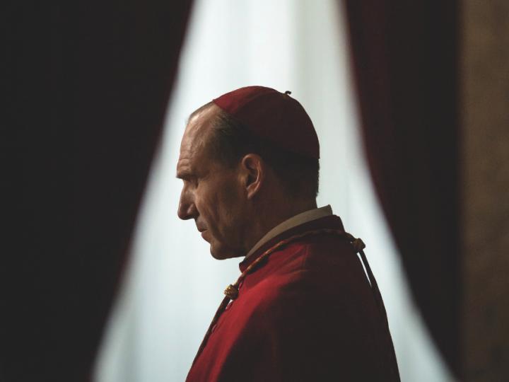 A white man in a red cap and red robe stands in front of a window. The perspective is from his side profile.