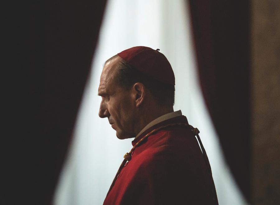 A white man in a red cap and red robe stands in front of a window. The perspective is from his side profile.