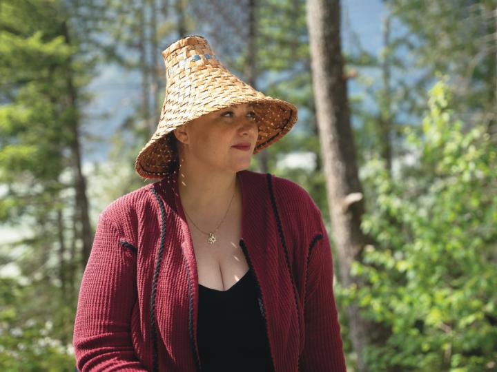 An Indigenous woman is wearing a traditional woven hate, a red coat and black shirt