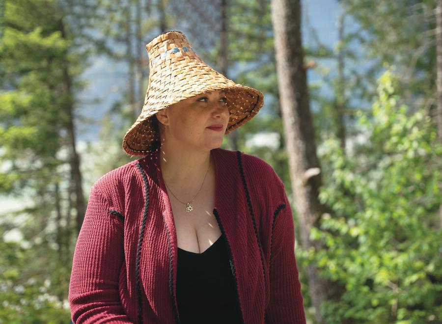 An Indigenous woman is wearing a traditional woven hate, a red coat and black shirt