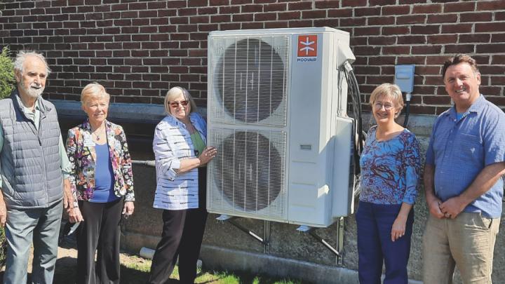 Five people stand in front of a building next to a heat pump. The heat pump is in the middle. Starting from the left is an older white man with grey hair. He is wearing a button up shirt and jeans with a navy vest. Next to him is an older woman with short white hair in a blue shirt and a floral blazer and black pants. Next to her is an older white woman with white hair, brown sunglasses and a striped white and black shirt with black pants. Her right hand rests on the white heat pump, which has two vertical fans. Next to the heat pump is an older white woman with white hair, glasses, a floral top and black pants. Next to her is a middle-aged white man with brown hair, blue shirt and beige slacks.