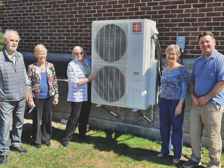 Five people stand in front of a building next to a heat pump. The heat pump is in the middle. Starting from the left is an older white man with grey hair. He is wearing a button up shirt and jeans with a navy vest. Next to him is an older woman with short white hair in a blue shirt and a floral blazer and black pants. Next to her is an older white woman with white hair, brown sunglasses and a striped white and black shirt with black pants. Her right hand rests on the white heat pump, which has two vertical fans. Next to the heat pump is an older white woman with white hair, glasses, a floral top and black pants. Next to her is a middle-aged white man with brown hair, blue shirt and beige slacks.